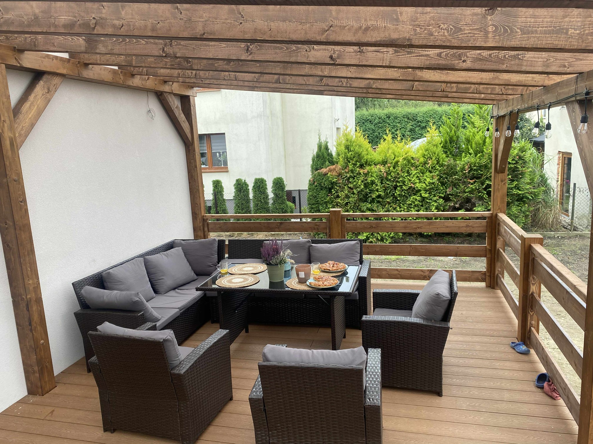 Drewniany taras z zadaszeniem i meblami ogrodowymi. Zastawiony stół z pizzą i napojami. W tle budynek i zieleń. Przytulna aranżacja przestrzeni.