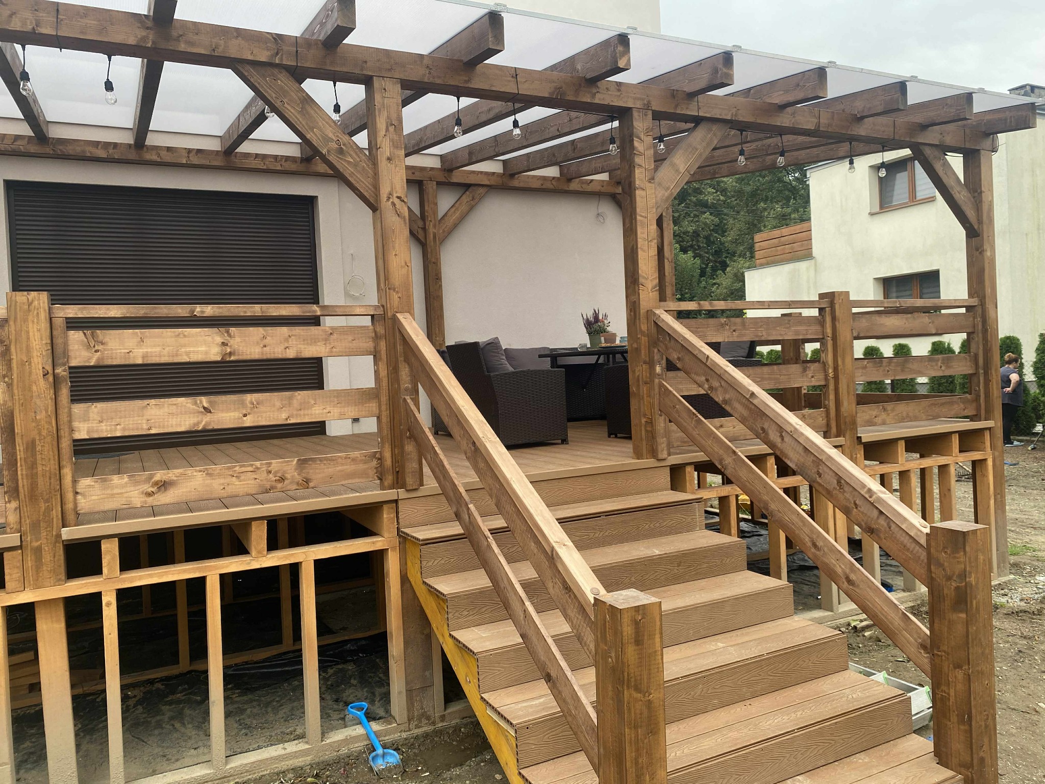 Drewniany taras z zadaszeniem w trakcie budowy, widoczne schody i konstrukcja, oświetlenie w postaci żarówek, niebieska łopatka leży obok schodów.
