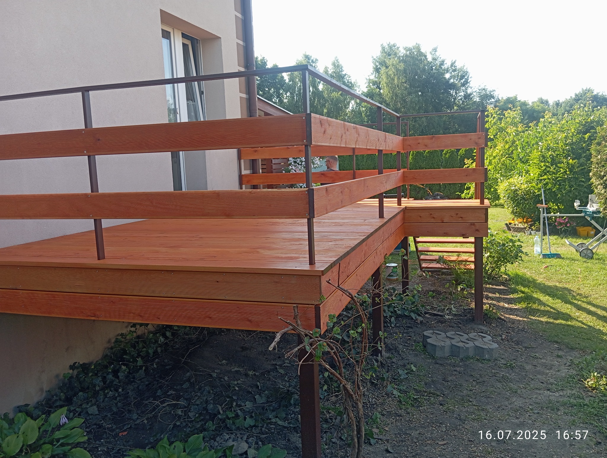 Nowoczesny taras drewniany z brązowymi balustradami i schodami, przylegający do domu. Widok na zadbany ogród w tle, zielona trawa i drzewa. Słoneczny dzień.