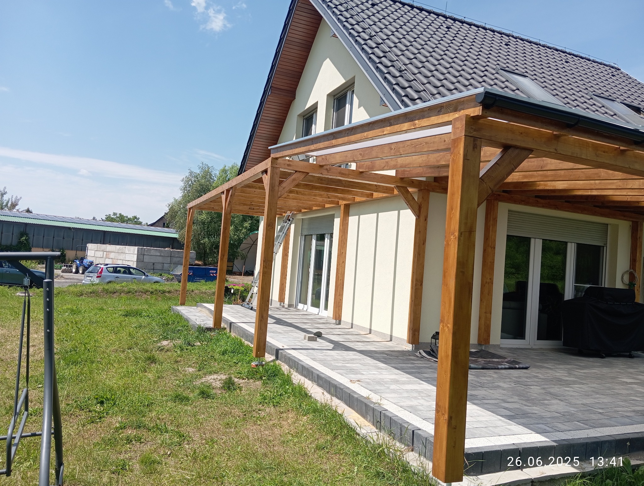 Drewniana pergola z transparentnym zadaszeniem, przylegająca do jasnego budynku z szarą dachówką. Widoczny fragment tarasu wyłożonego kostką brukową i zielony trawnik.