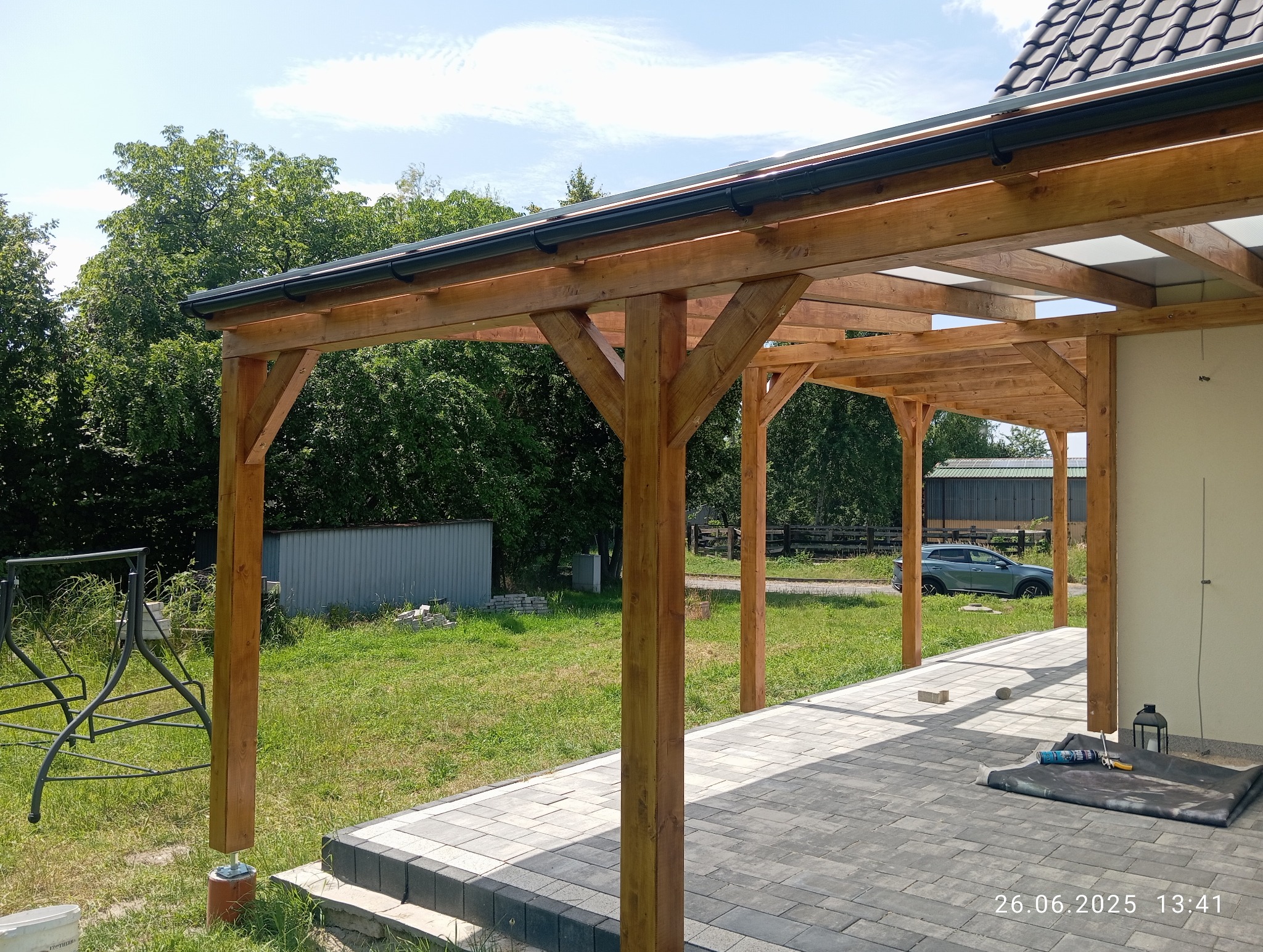 Drewniana pergola z czarną rynną, częściowo zadaszona, na tle zieleni i kostki brukowej. Widoczne narzędzia i data. Słoneczny dzień.