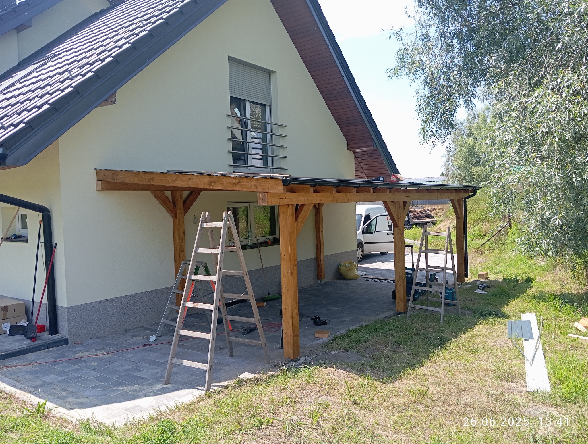 Drewniana konstrukcja z zadaszeniem przy domu, widoczne drabiny i narzędzia, sugerujące trwający montaż. Jasna elewacja budynku z oknem i roletą.