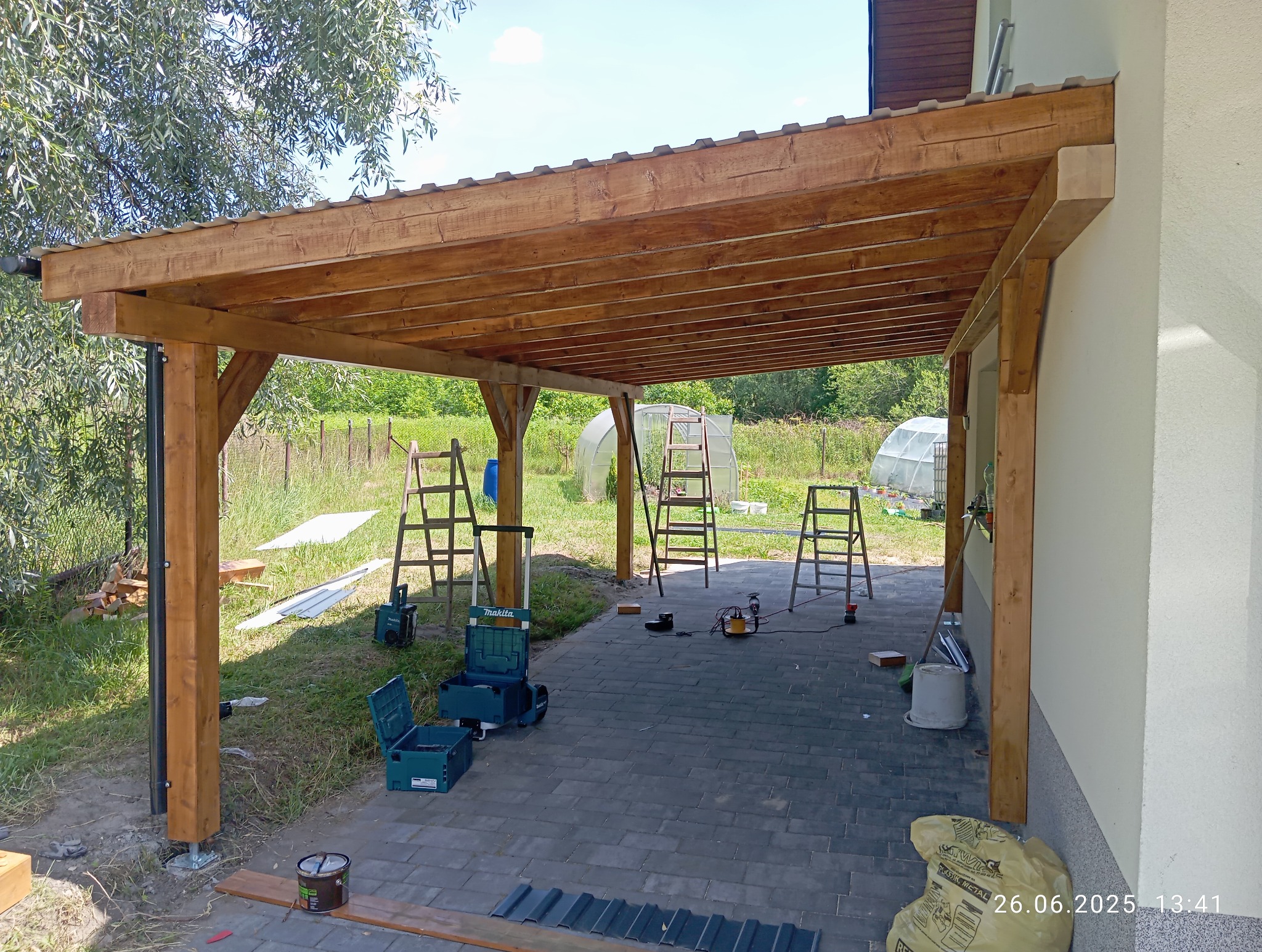 Drewniana konstrukcja z zadaszeniem, widoczne narzędzia i drabiny, sugerujące etap budowy. W tle zieleń i szklarnie. Elementy montażowe i wykończeniowe na ziemi.