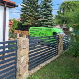 Nowoczesne ogrodzenie panelowe w kolorze grafitowym z kamiennymi słupkami, na tle zielonego firmowego busa i zabudowań.