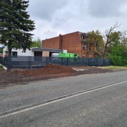 Werka Bramy Weronika Wanczura - Nowoczesne ogrodzenie panelowe w kolorze grafitowym, oddzielające posesję z białym domem od drogi, z widoczną stertą ziemi i zielonym busem w tle.