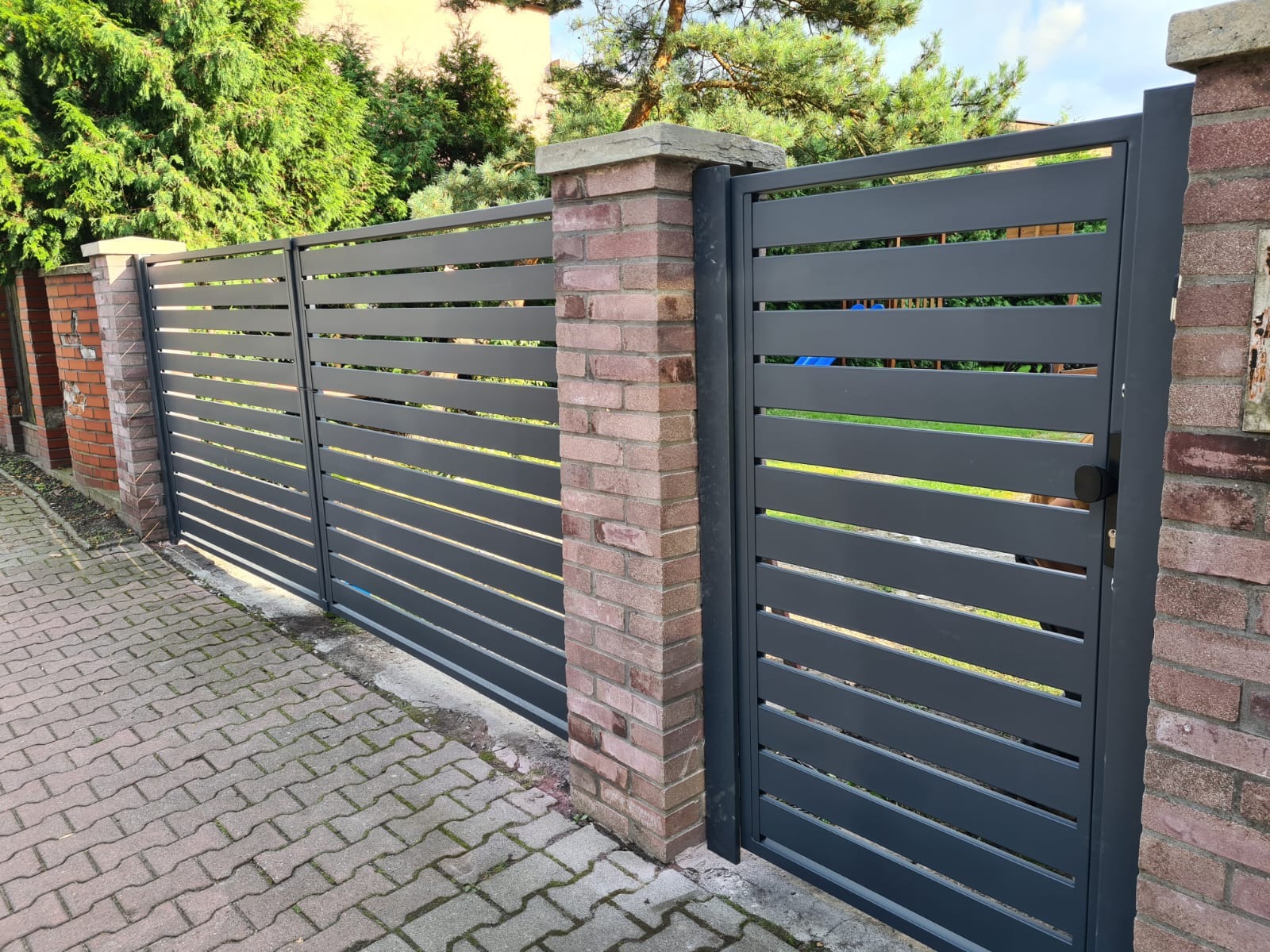 Nowoczesna, grafitowa brama wjazdowa z poziomymi panelami, osadzona na ceglanych słupkach z betonowymi daszkami, widok z zewnątrz na kostkę brukową i zieleń ogrodu.