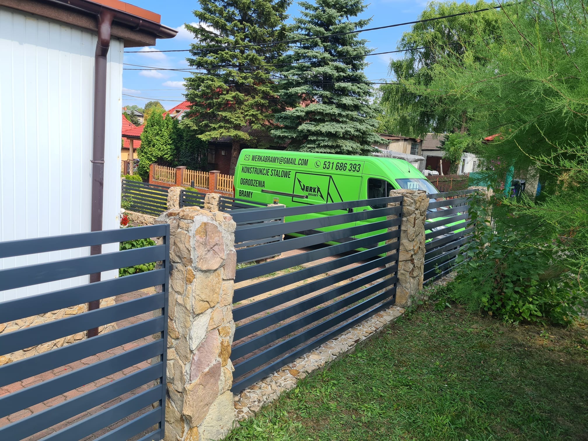 Nowoczesne ogrodzenie panelowe w kolorze grafitowym z kamiennymi słupkami, na tle zielonego firmowego busa i zabudowań.