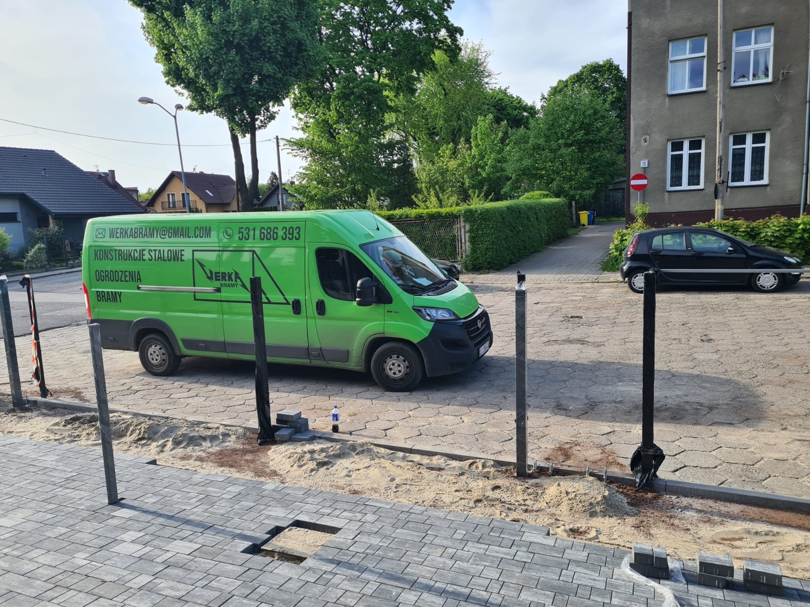 Montaż słupków ogrodzeniowych na posesji, widoczny firmowy zielony bus z danymi kontaktowymi i logo firmy.
