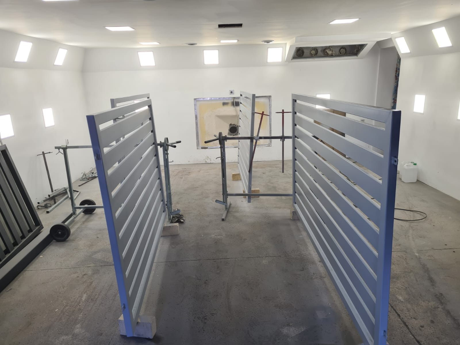 Elementy bramy wjazdowej z poziomymi szczeblami, świeżo pomalowane, ustawione w lakierni na metalowych stojakach, widoczne ślady po lakierowaniu na podłodze.