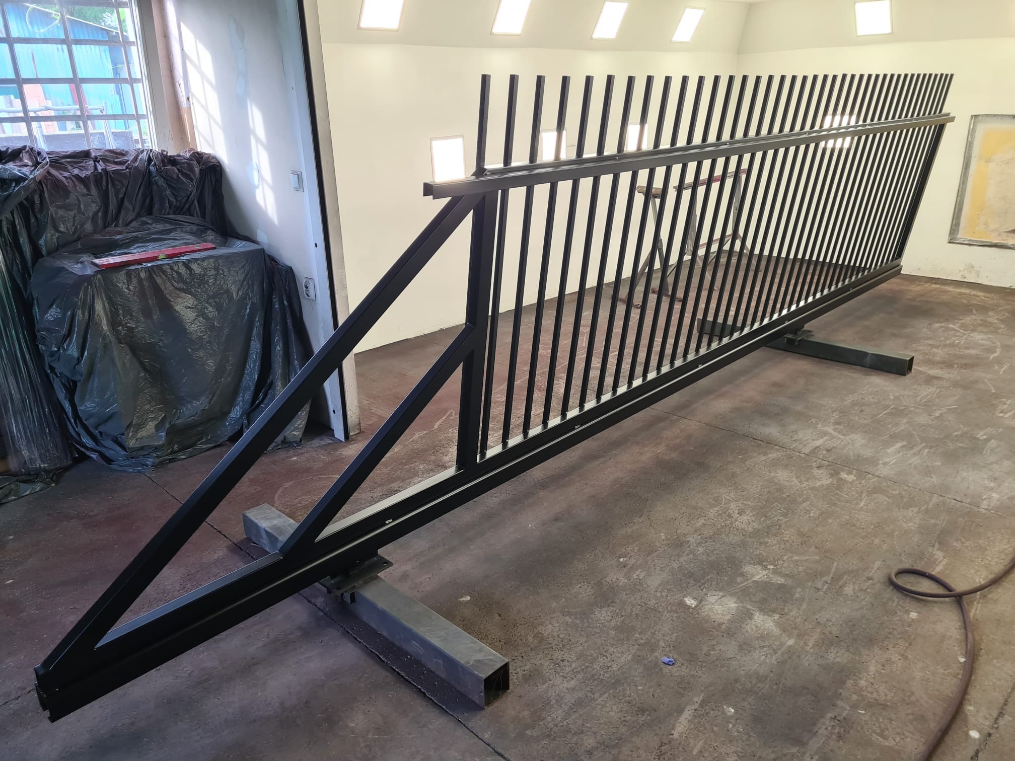 Surowa, czarna, metalowa konstrukcja bramy na metalowych wspornikach w warsztacie, widoczna poziomica na przykrytym folią stole, ślady farby na podłodze.