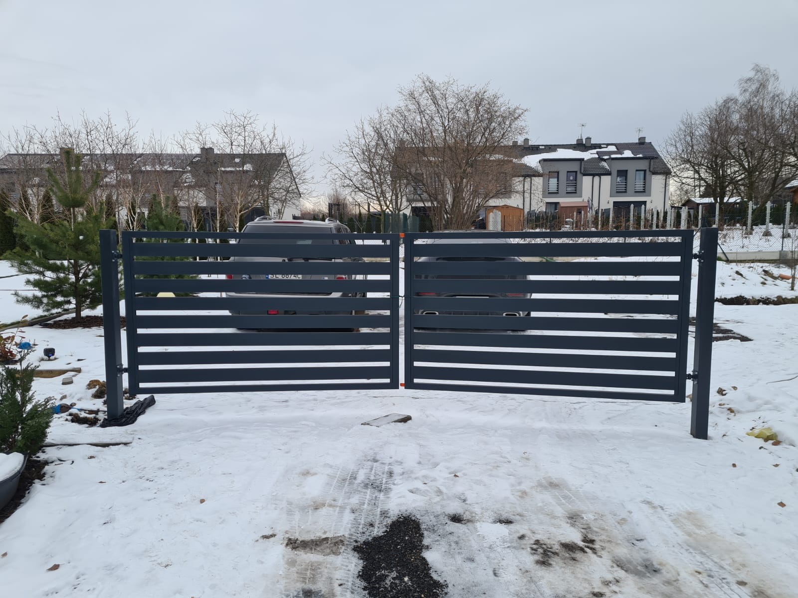 Nowoczesna, pozioma brama wjazdowa w kolorze antracytowym, widok z zewnątrz na posesję z zaparkowanym samochodem w zimowej scenerii.
