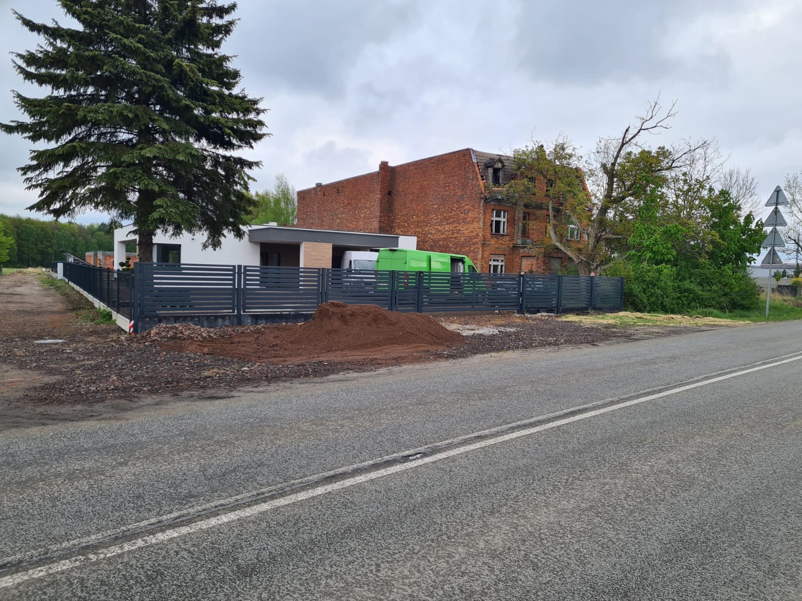 Nowoczesne ogrodzenie panelowe w kolorze grafitowym, oddzielające posesję z białym domem od drogi, z widoczną stertą ziemi i zielonym busem w tle.