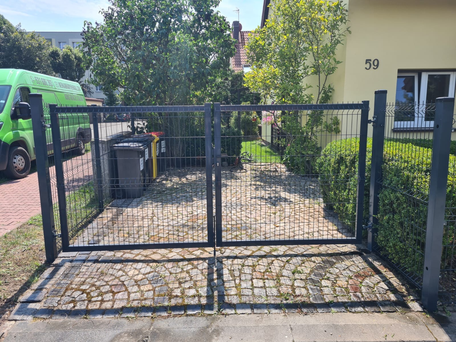 Metalowa, dwuskrzydłowa brama wjazdowa z siatki na posesję z brukowanym podjazdem, widok z zewnątrz, na tle zieleni i domu z numerem 59.