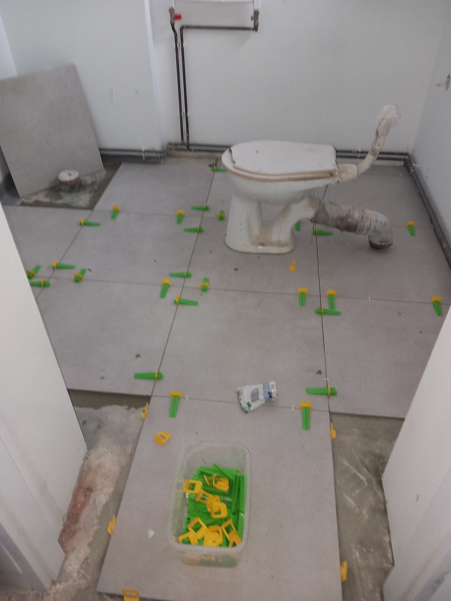 Szare płytki podłogowe w trakcie układania w łazience z widoczną toaletą i poziomującymi klipsami oraz pojemnikiem z klinami.