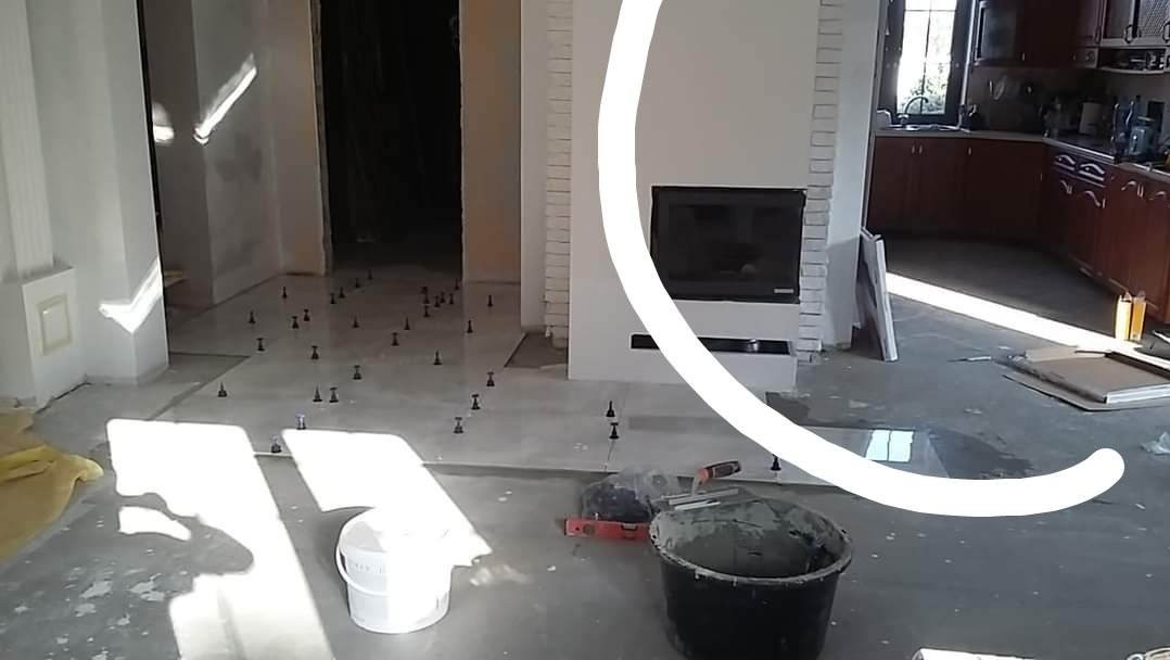 Wnętrze w trakcie prac wykończeniowych: przygotowywana podłoga pod płytki, widoczne poziomica, wiadro z zaprawą, krzyżyki dystansowe, w tle kominek i zabudowa kuchenna.