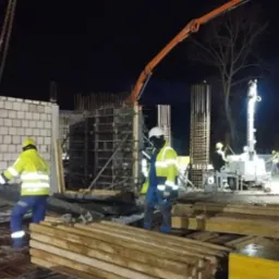 Prace budowlane w nocy: robotnicy w odblaskowych ubraniach układają szalunki na zbrojeniu fundamentów, w tle widoczny dźwig z pompą do betonu i ściana z białej cegły.