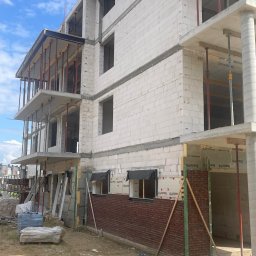 DD FIRMA USŁUGOWO-BUDOWLANA - Wznoszenie wielopiętrowego budynku z pustaków, częściowo obłożonego czerwoną cegłą, z widocznymi elementami konstrukcyjnymi i rusztowaniami, na tle błękitnego nieba.