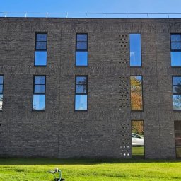 DD FIRMA USŁUGOWO-BUDOWLANA - Dwupiętrowy budynek z ciemnej cegły z regularnie rozmieszczonymi oknami, na trawniku widoczny hydrant i zaparkowany samochód.