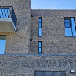 DD FIRMA USŁUGOWO-BUDOWLANA - Nowoczesny, dwupiętrowy dom z ciemnej cegły z balkonem i dużymi oknami w Działdowie, widziany z dołu pod kątem.