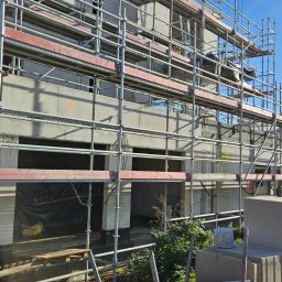 DD FIRMA USŁUGOWO-BUDOWLANA - Wznoszenie surowego, betonowego budynku wielorodzinnego z użyciem rusztowań, widoczne bloki betonu komórkowego i fragment twarzy osoby w kasku.