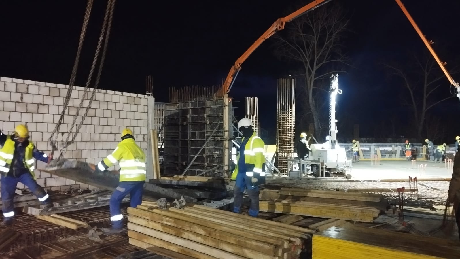 Prace budowlane w nocy: robotnicy w odblaskowych ubraniach układają szalunki na zbrojeniu fundamentów, w tle widoczny dźwig z pompą do betonu i ściana z białej cegły.