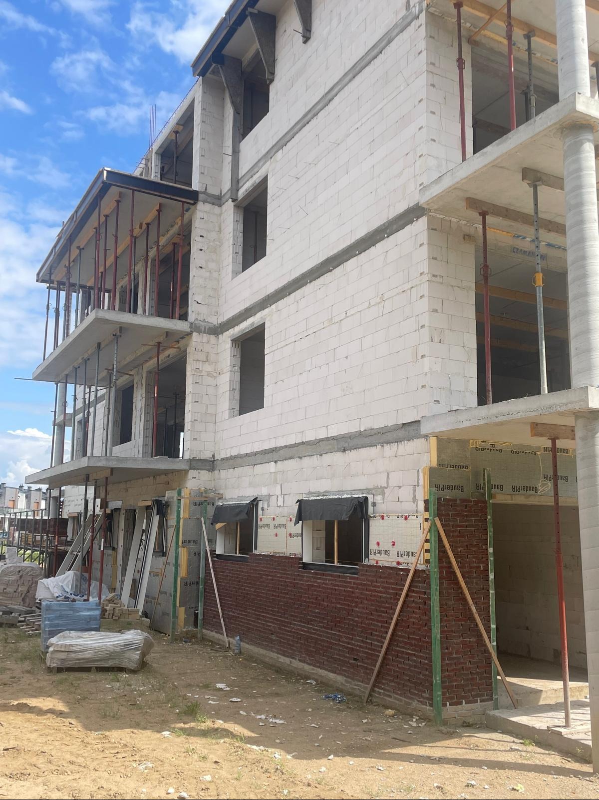 Wznoszenie wielopiętrowego budynku z pustaków, częściowo obłożonego czerwoną cegłą, z widocznymi elementami konstrukcyjnymi i rusztowaniami, na tle błękitnego nieba.