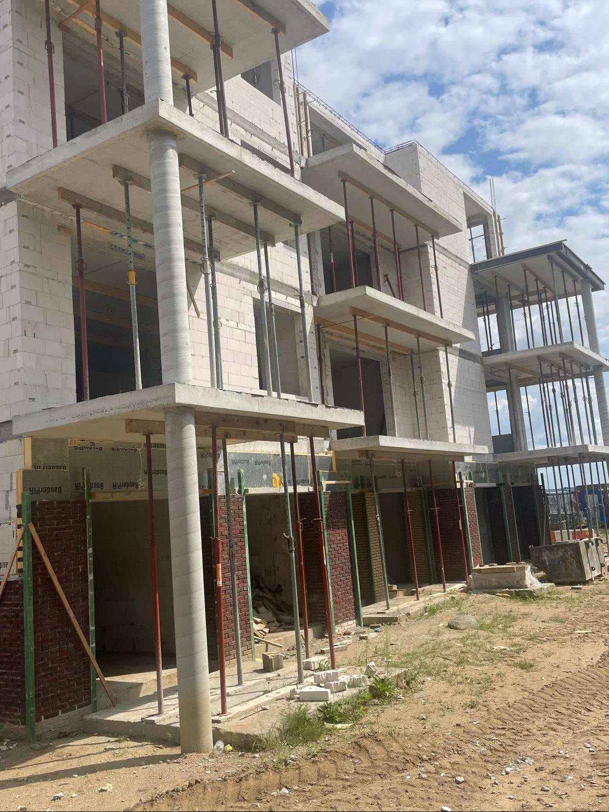 Wznoszenie wielopiętrowego budynku z białych bloczków, z widocznymi podporami stropów i fragmentami muru z cegły klinkierowej na parterze, w słoneczny dzień.