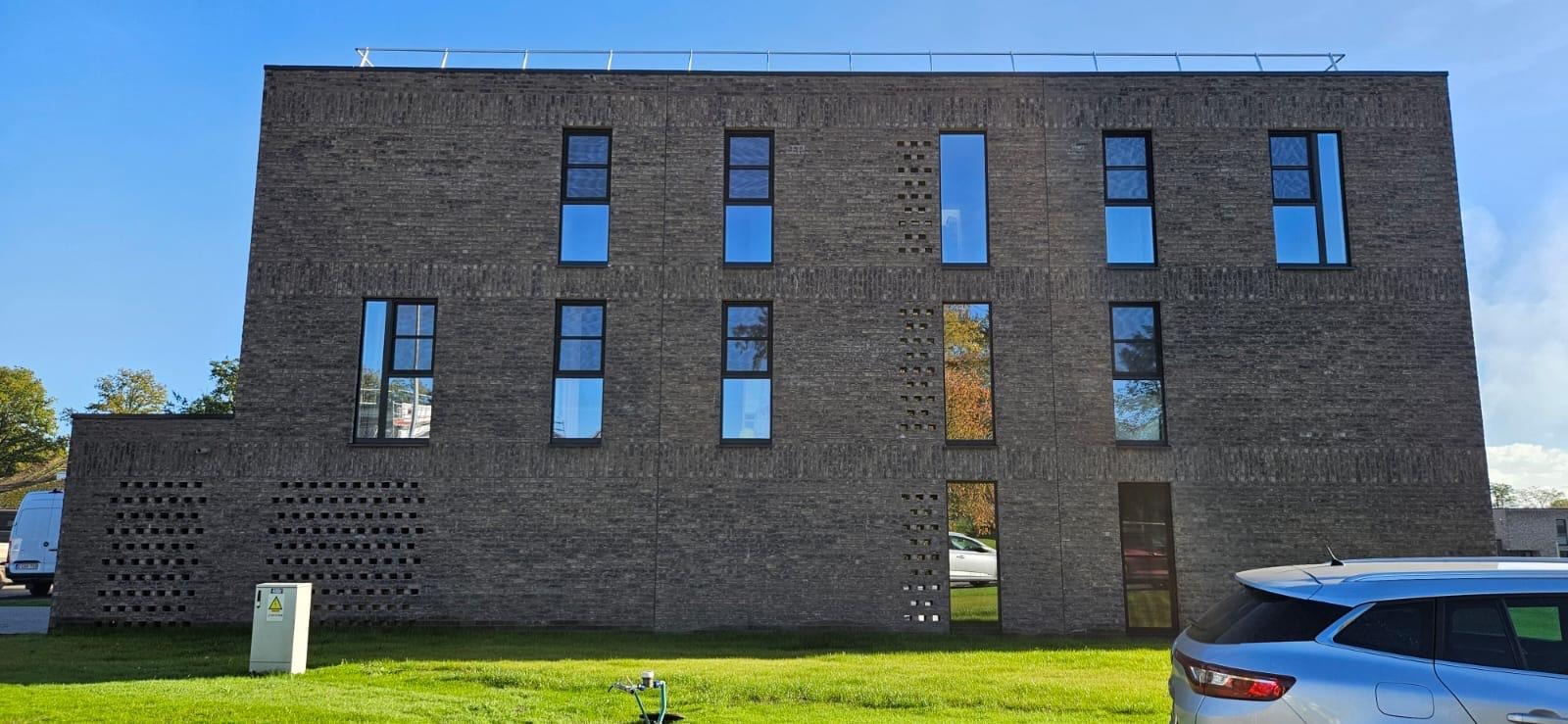 Dwupiętrowy budynek z ciemnej cegły z regularnie rozmieszczonymi oknami, na trawniku widoczny hydrant i zaparkowany samochód.