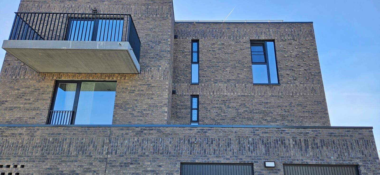 Nowoczesny, dwupiętrowy dom z ciemnej cegły z balkonem i dużymi oknami w Działdowie, widziany z dołu pod kątem.