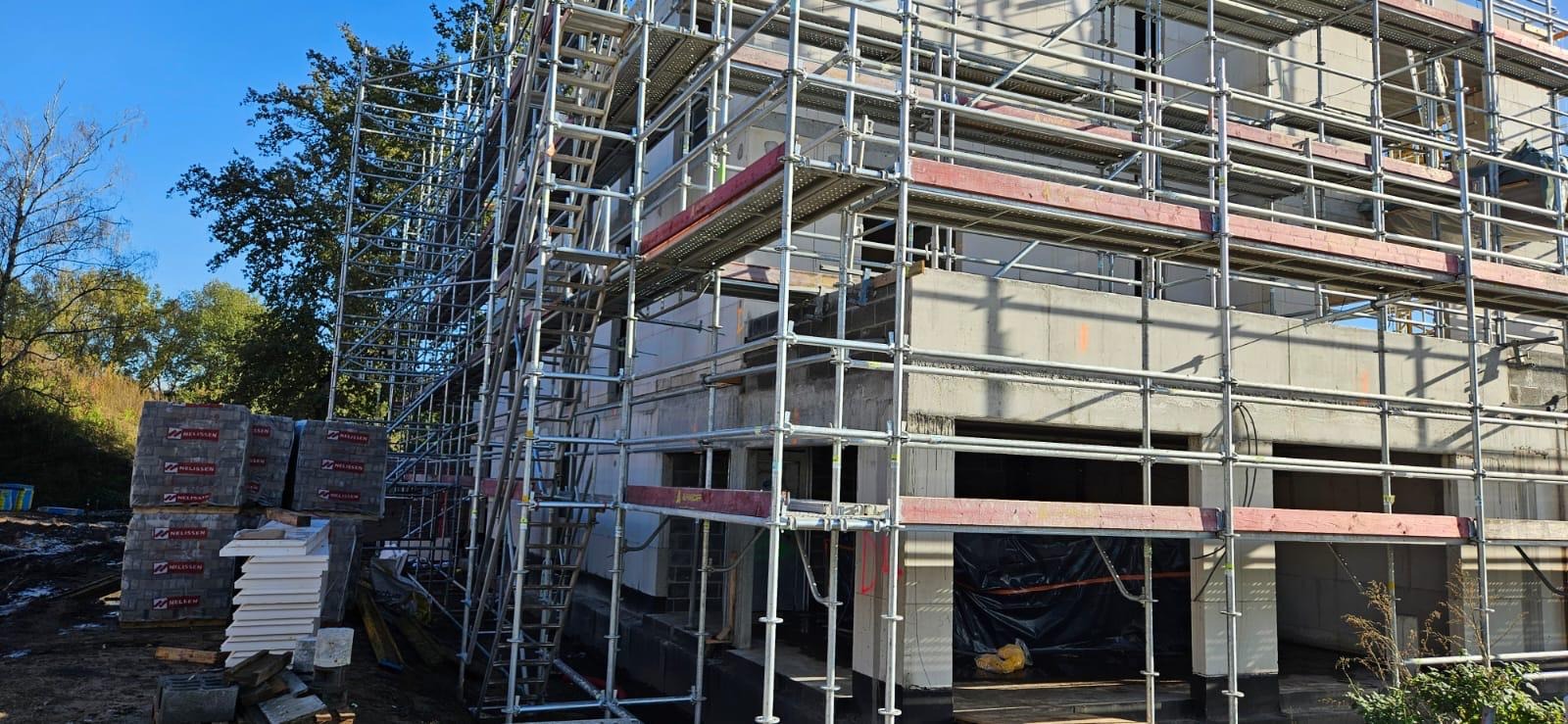 Wznoszony dwukondygnacyjny budynek w surowym stanie, otoczony rusztowaniami, z widocznymi stertami materiałów budowlanych na ziemi.