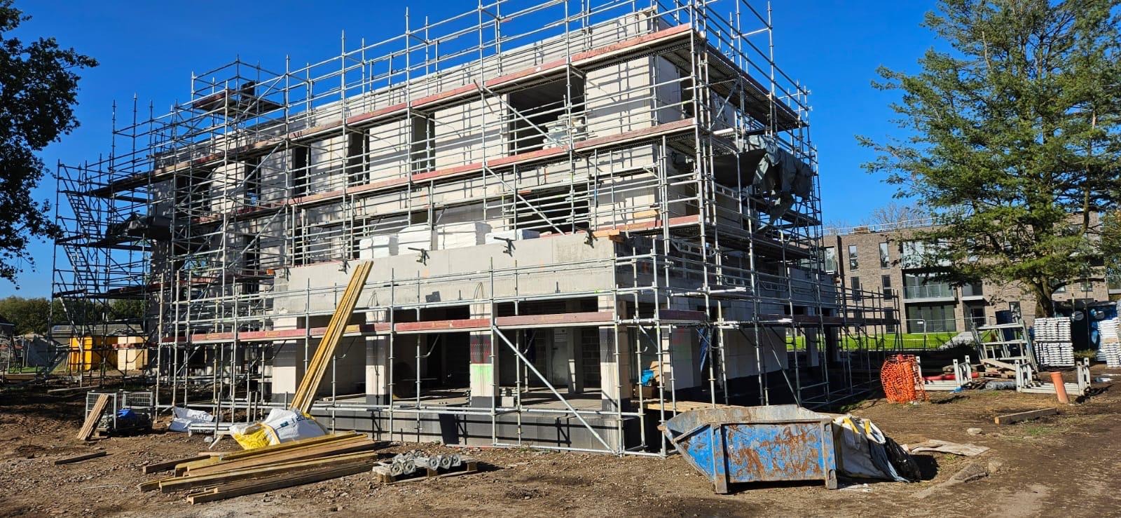 Surowy stan dwupiętrowego domu w trakcie budowy, otoczonego rusztowaniami, z widocznymi bloczkami betonowymi i elementami konstrukcyjnymi, na pierwszym planie deski i kontener na gruz, w tle drzewa...