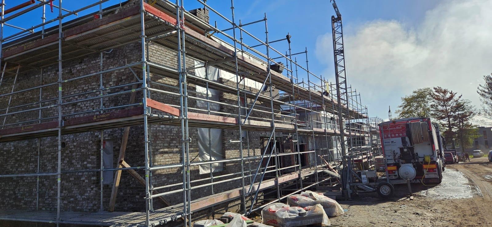 Budynek z cegły w trakcie prac budowlanych, otoczony rusztowaniami, z widocznym dźwigiem i samochodem dostawczym firmy Carro-Bel w tle.