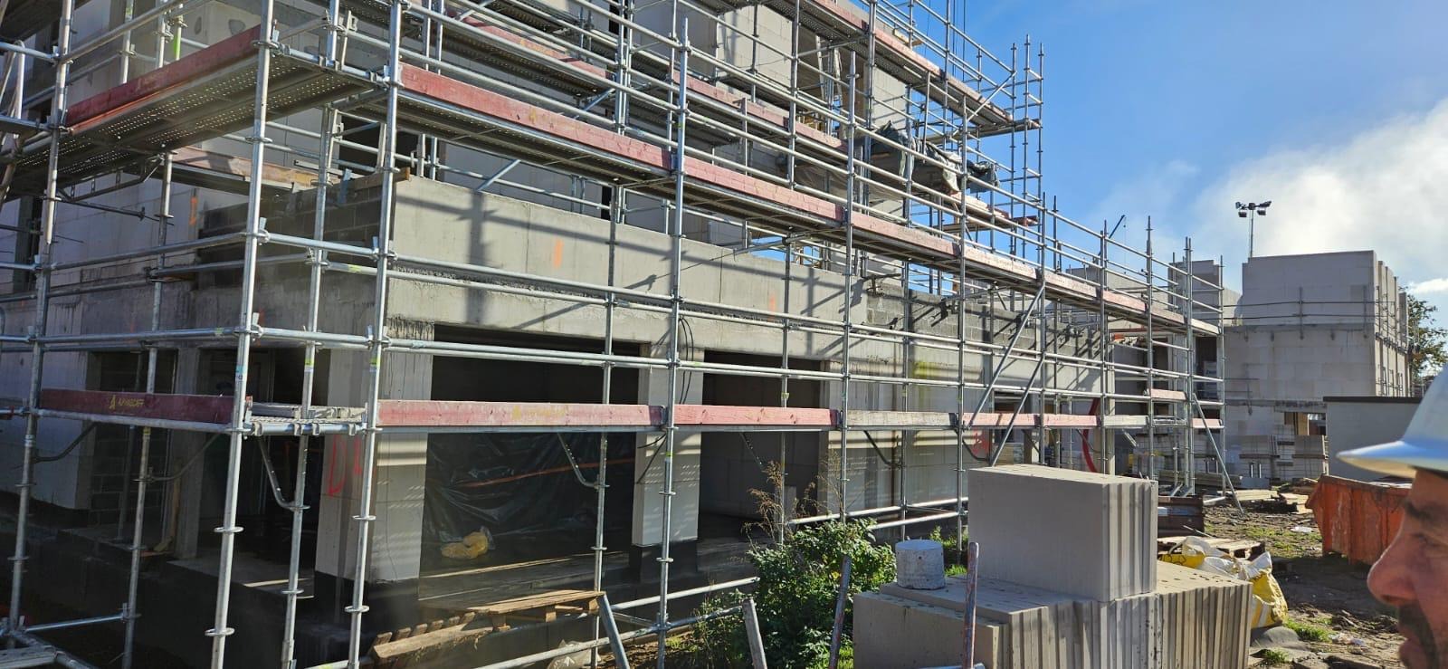 Wznoszenie surowego, betonowego budynku wielorodzinnego z użyciem rusztowań, widoczne bloki betonu komórkowego i fragment twarzy osoby w kasku.