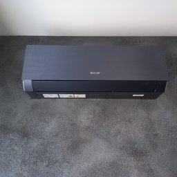 GreenVents KONRAD KOŹBIAŁ - Czarny klimatyzator Sinclair zamontowany na ścianie o grafitowej strukturze betonu. Widok z dołu, podkreślający nowoczesny design urządzenia i jego wkomponowanie w wystrój.