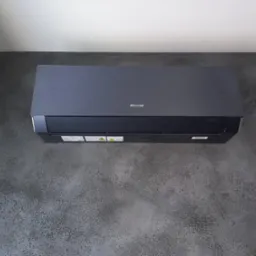 Czarny klimatyzator Sinclair zamontowany na ścianie o grafitowej strukturze betonu. Widok z dołu, podkreślający nowoczesny design urządzenia i jego wkomponowanie w wystrój.