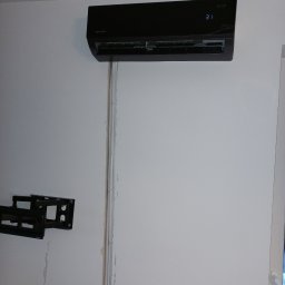 GreenVents KONRAD KOŹBIAŁ - Czarny klimatyzator Rotenso Elis zamontowany na białej ścianie, widoczne przewody w listwach. Na ścianie uchwyt do TV.