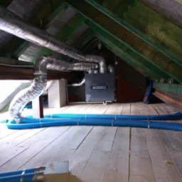 System wentylacji z rekuperacją w przestrzeni poddasza z drewnianą konstrukcją. Widoczne srebrne rury wentylacyjne i niebieskie przewody elastyczne.