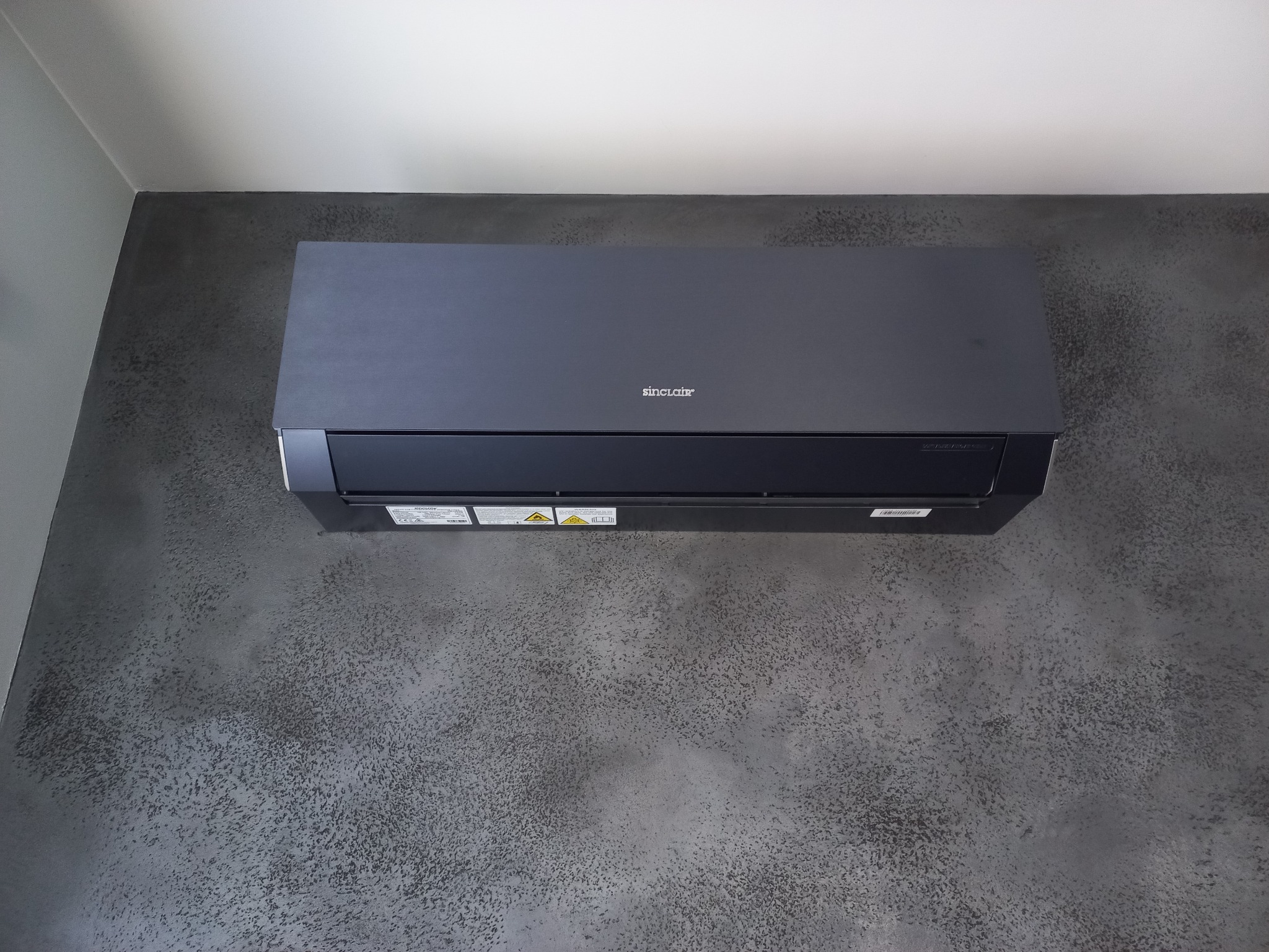 Czarny klimatyzator Sinclair zamontowany na ścianie o grafitowej strukturze betonu. Widok z dołu, podkreślający nowoczesny design urządzenia i jego wkomponowanie w wystrój.