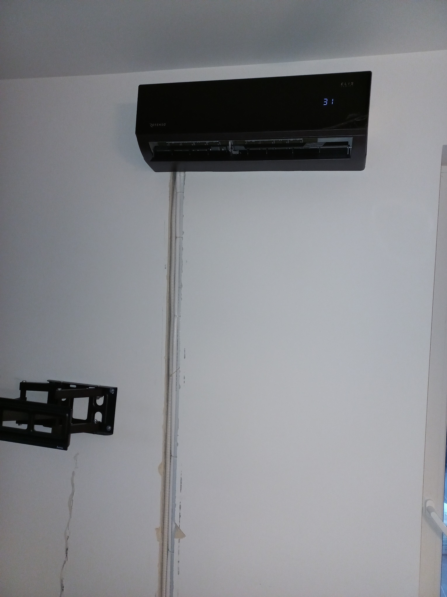 Czarny klimatyzator Rotenso Elis zamontowany na białej ścianie, widoczne przewody w listwach. Na ścianie uchwyt do TV.