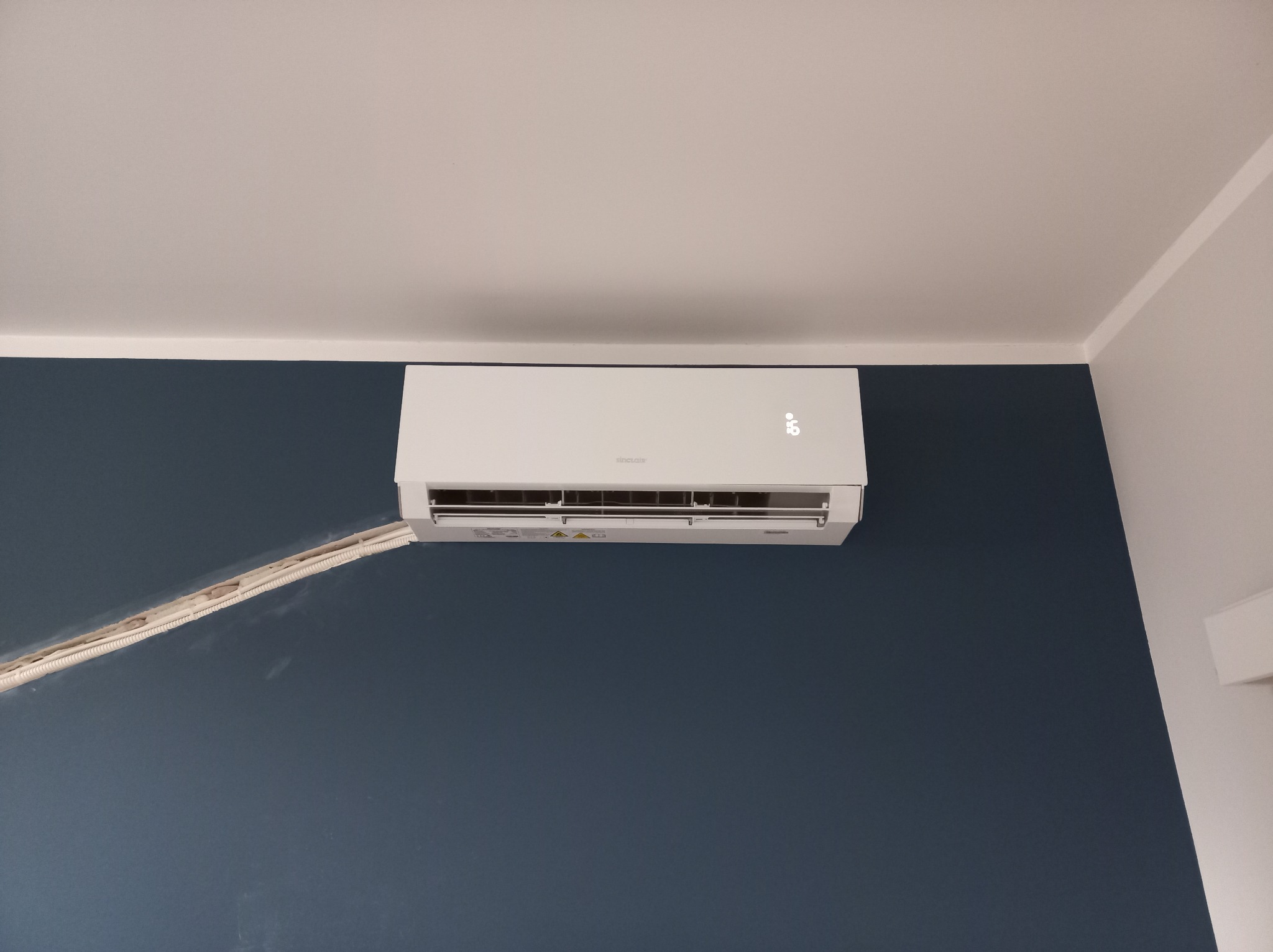 Biały klimatyzator Sinclair na granatowej ścianie, widoczne kable w białej listwie. Minimalistyczny design, temperatura 16 stopni Celsjusza.