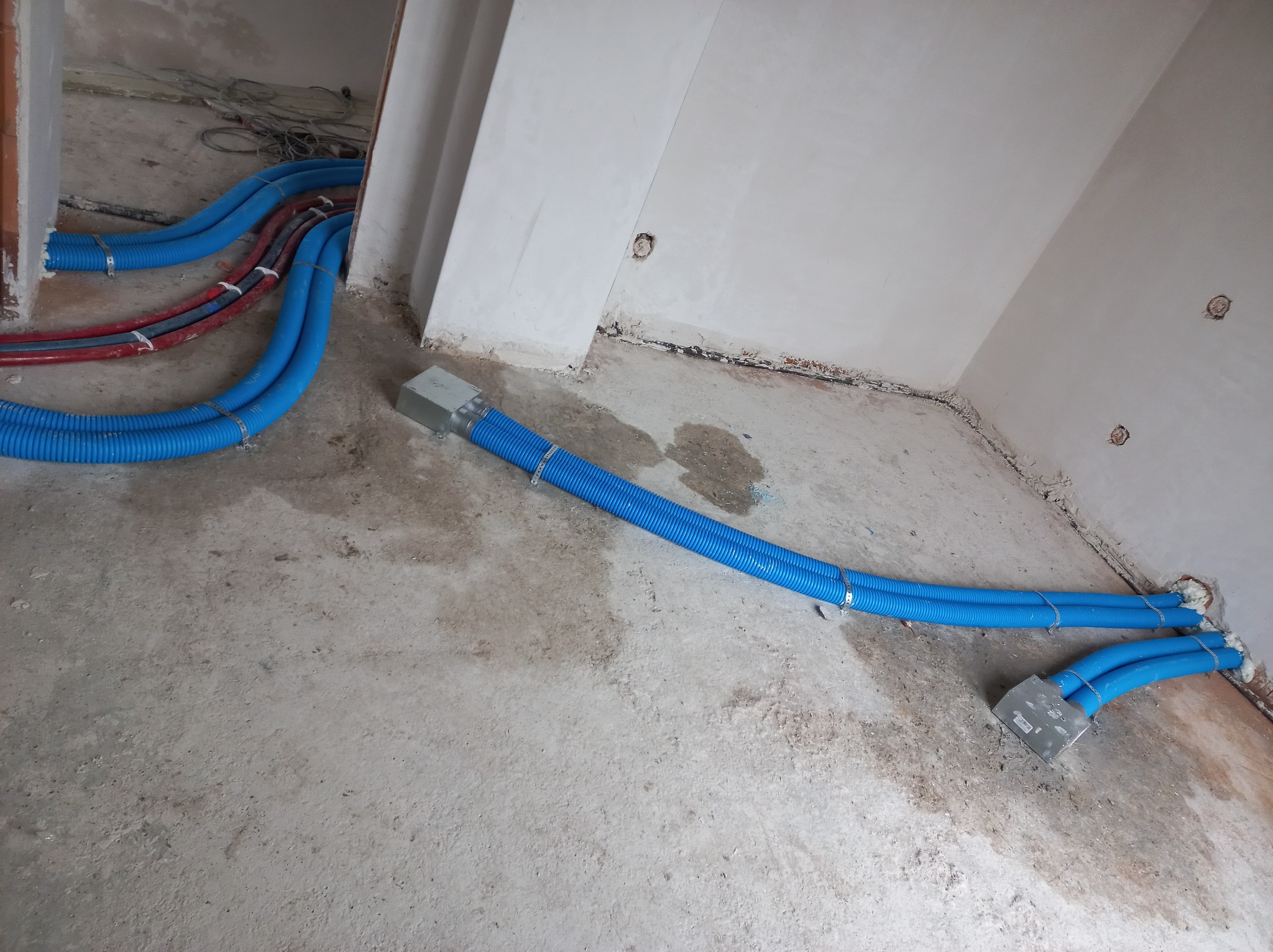 Niebieskie i czerwone rury wentylacyjne, częściowo zakryte, prowadzone po betonowej podłodze w surowym wnętrzu budynku w trakcie instalacji.
