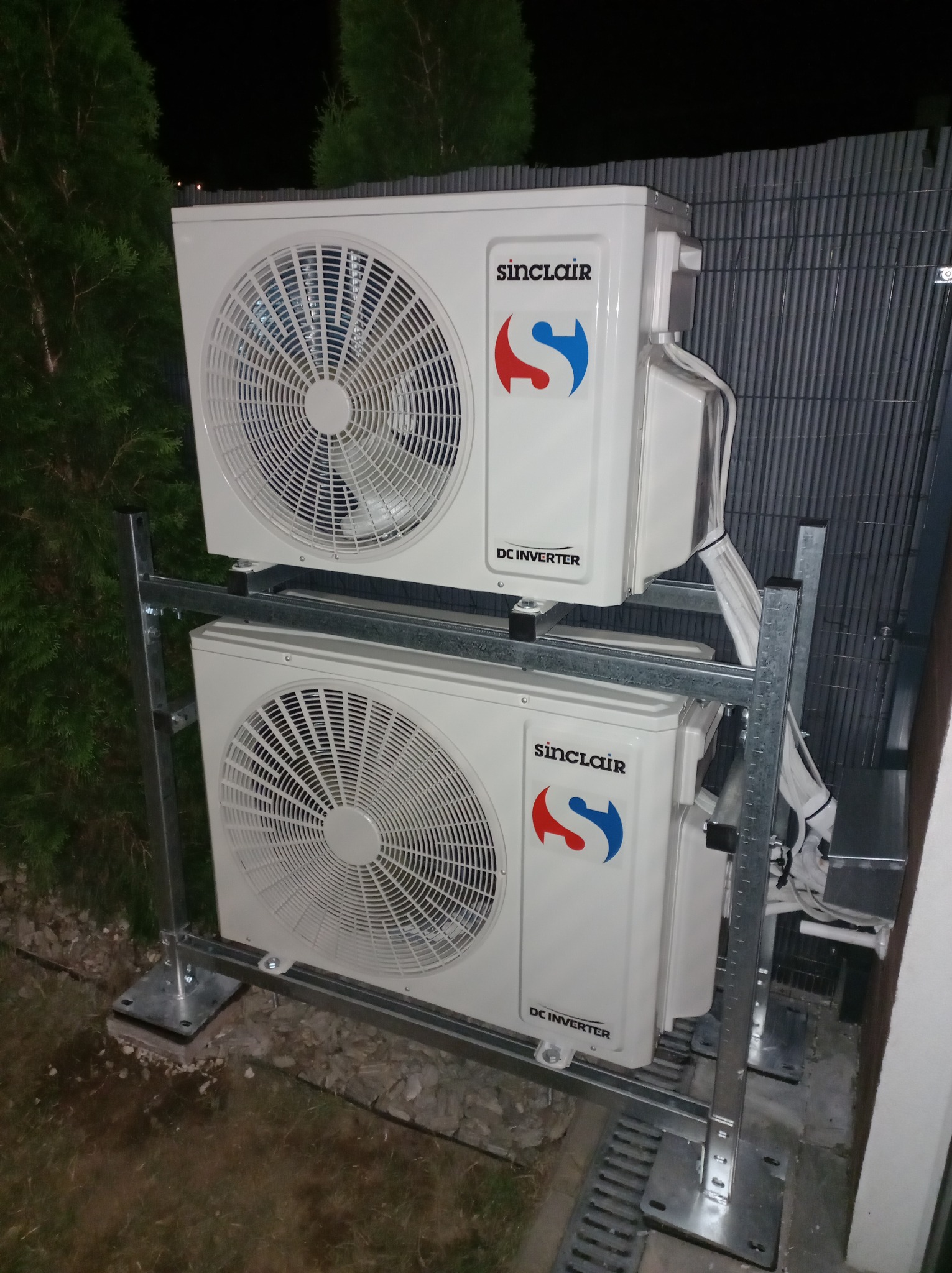 Dwa klimatyzatory Sinclair DC Inverter zamontowane jeden nad drugim na metalowej konstrukcji, widoczne przewody i elementy montażowe, tło stanowi ciemna zieleń i szara siatka.