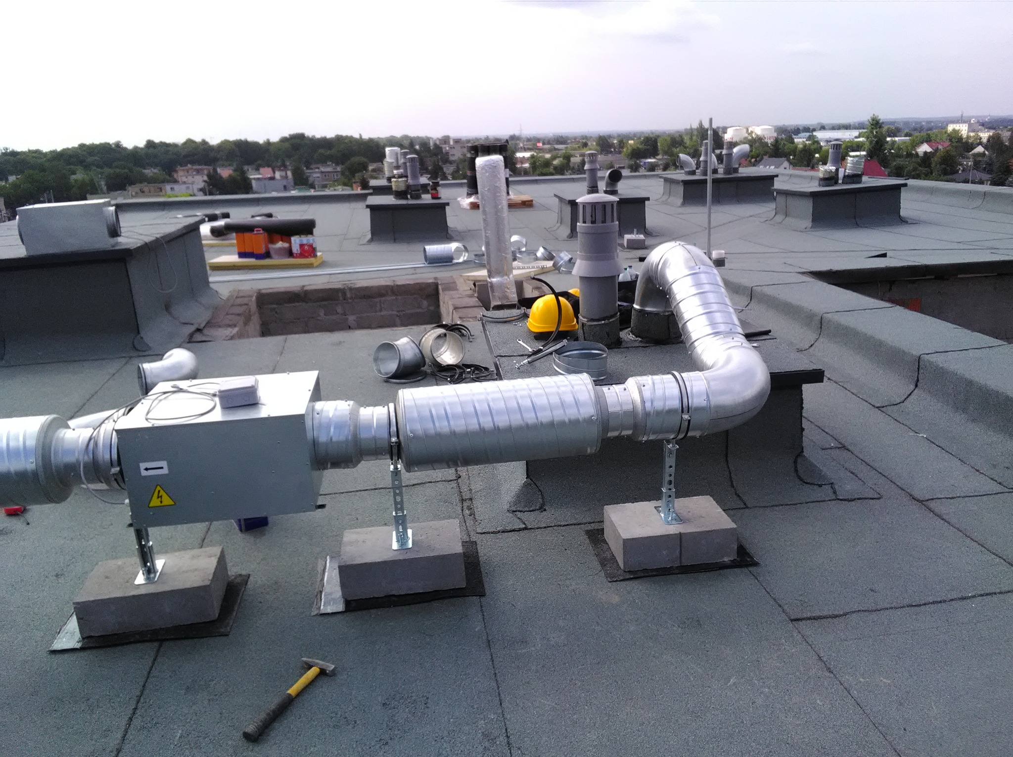 Instalacja wentylacyjna na dachu budynku z widocznymi przewodami, elementami montażowymi i narzędziami. W tle panorama miasta z zielenią.