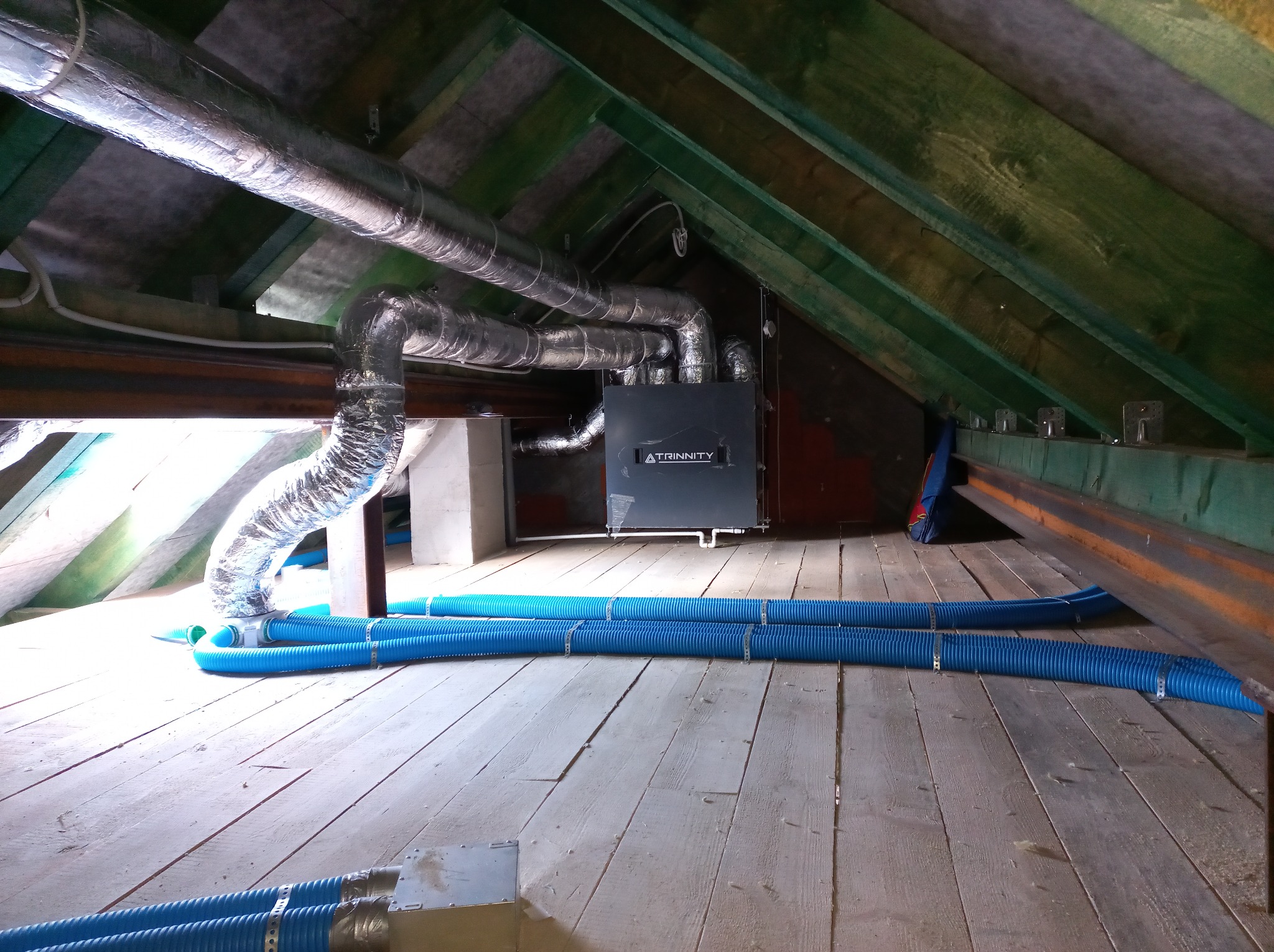 System wentylacji z rekuperacją w przestrzeni poddasza z drewnianą konstrukcją. Widoczne srebrne rury wentylacyjne i niebieskie przewody elastyczne.