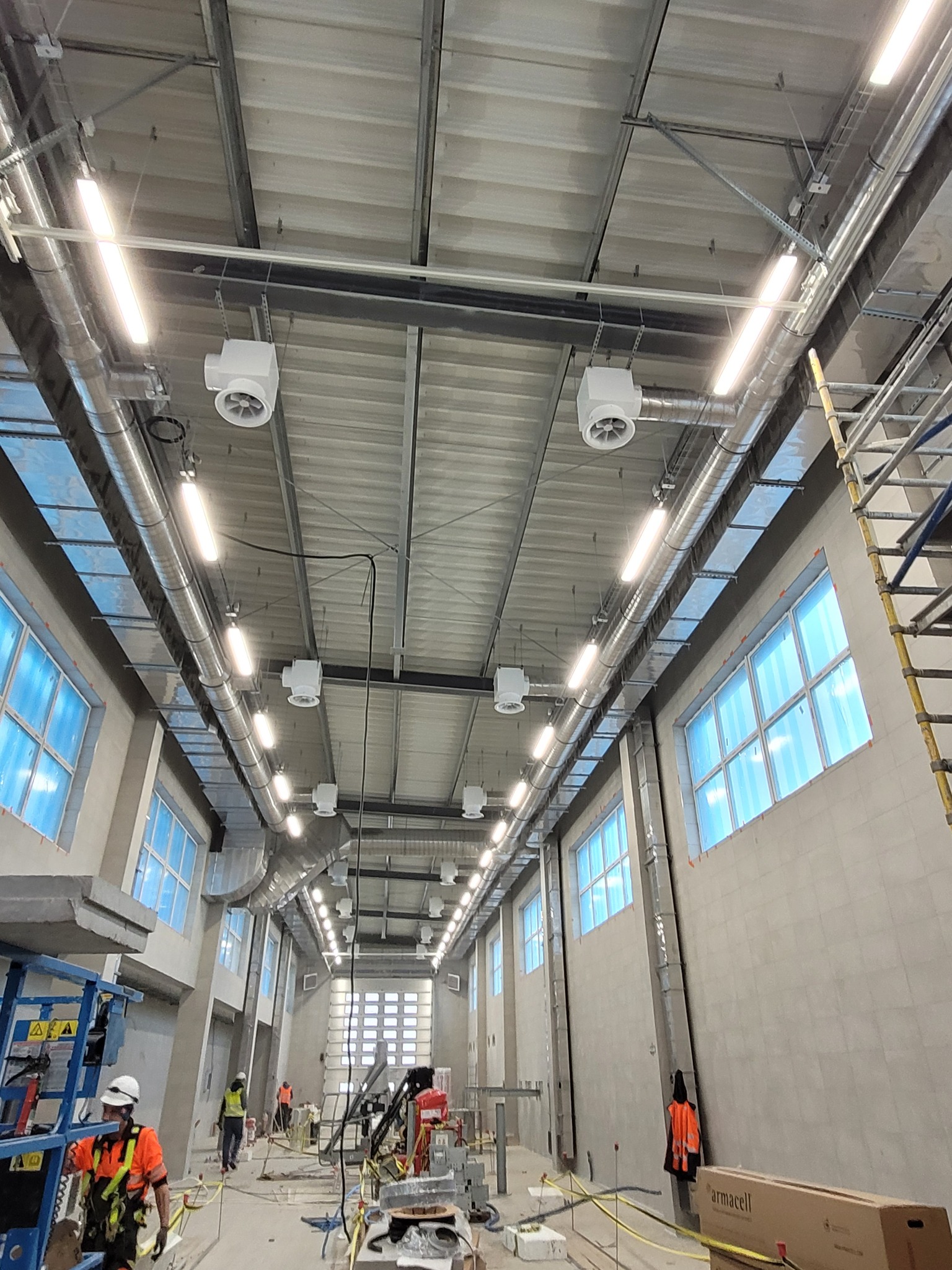 Wnętrze hali w trakcie montażu wentylacji: widoczne kanały wentylacyjne pod sufitem, białe nawiewniki, okna i pracownicy w odblaskowych ubraniach.