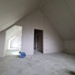 MIL-BUD SYLWESTER CHOMICZ - Poddasze z zamontowanymi płytami gipsowo-kartonowymi na ścianach i skosach. Widoczne otwory drzwiowe i narzędzia. Surowy stan wykończenia, brak szpachlowania.