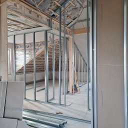 MIL-BUD SYLWESTER CHOMICZ - Montaż metalowej konstrukcji ścian działowych w surowym wnętrzu poddasza z izolacją termiczną. Widoczne profile, płyty i narzędzia.