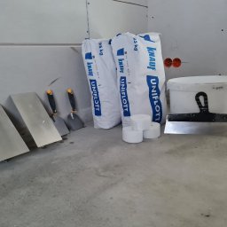 MIL-BUD SYLWESTER CHOMICZ - Przygotowane narzędzia i materiały budowlane: paka z masą szpachlową Knauf Uniflott, szpachelki i wiadro na tle ściany z płyt karton-gips, przed rozpoczęciem prac wykończeniowych.
