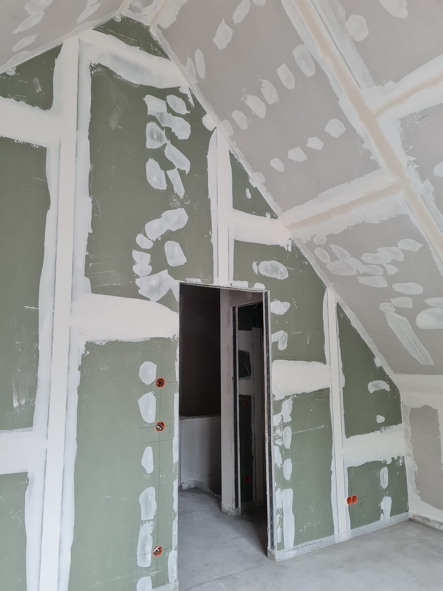 Poddasze w trakcie prac wykończeniowych. Widoczne płyty kartonowo-gipsowe na ścianach i suficie, częściowo pokryte masą szpachlową. Otwór drzwiowy w tle.