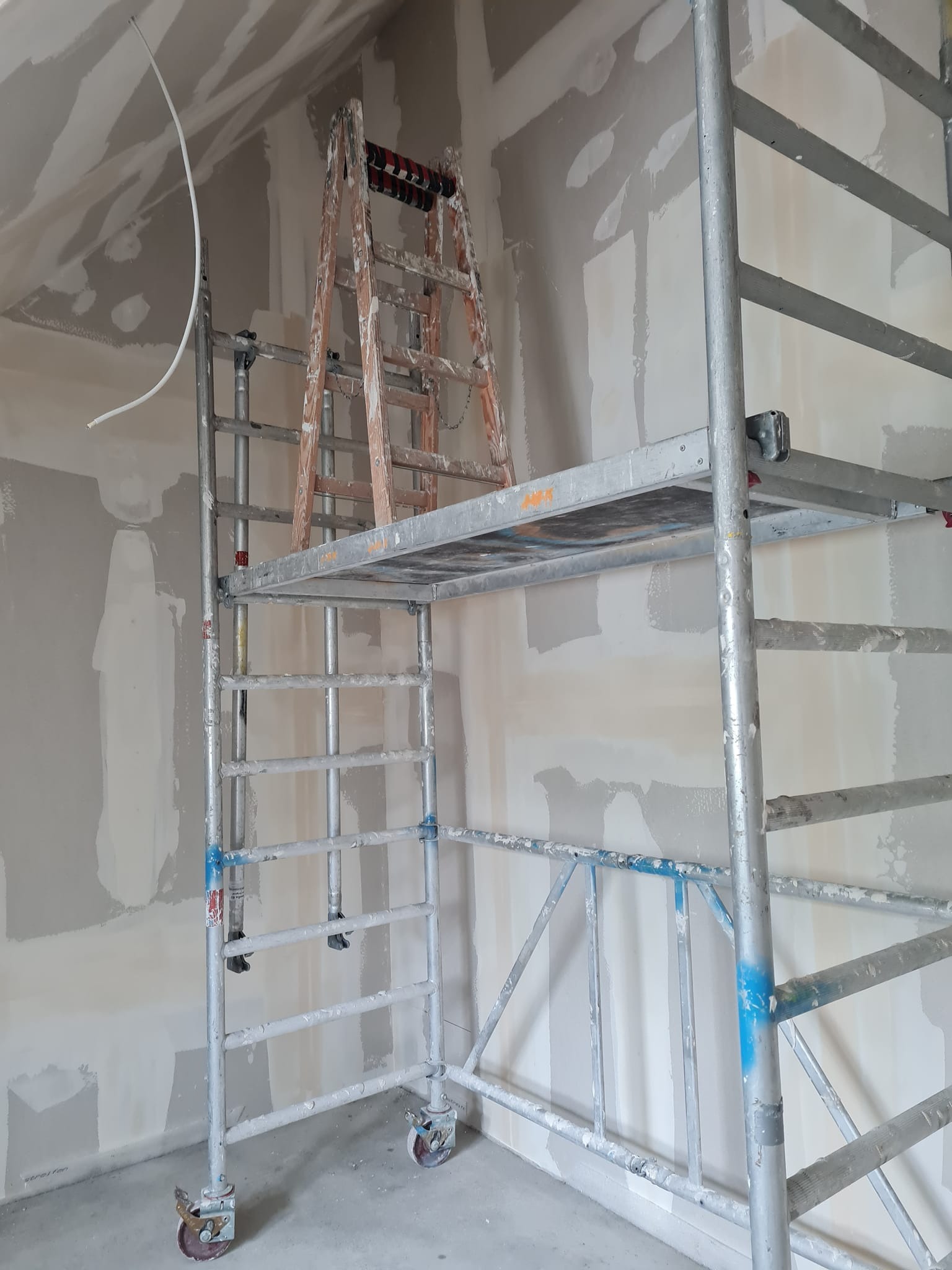 Wnętrze w trakcie prac wykończeniowych: ściany częściowo pokryte gładzią, widoczne rusztowanie aluminiowe i drewniana drabina oparta o ścianę. Przewód zwisa z sufitu.